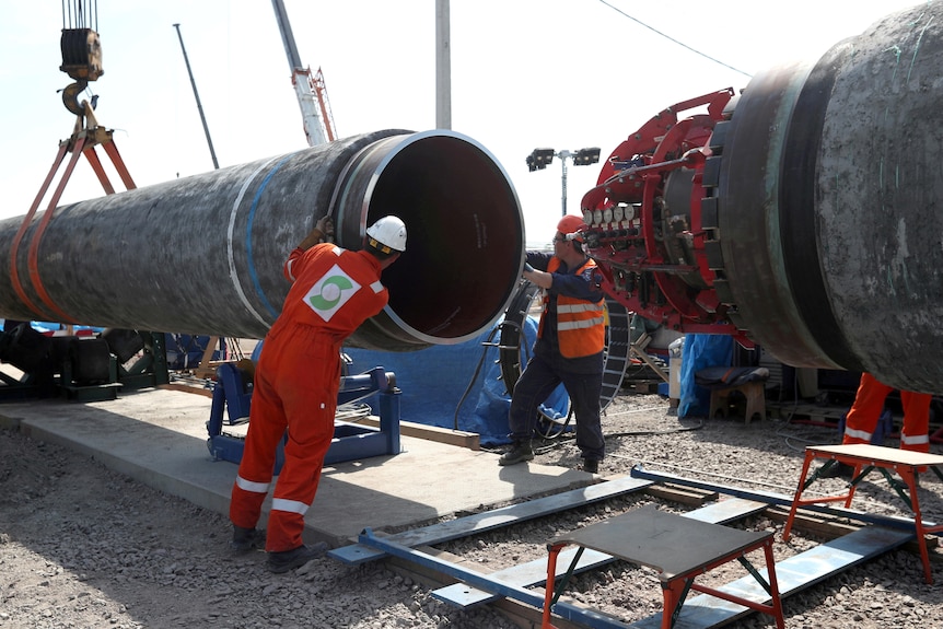 Des ouvriers stabilisent un gros tuyau métallique tenu par une grue.