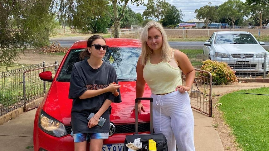 Two women stand in front of a red car.