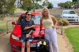 Two women stand in front of a red car.
