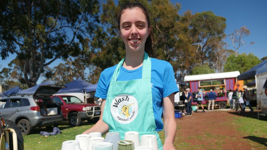 A teenager in a blue apron holding a tray of coffee mugs standing in front of food vendors at the local farmers' market.