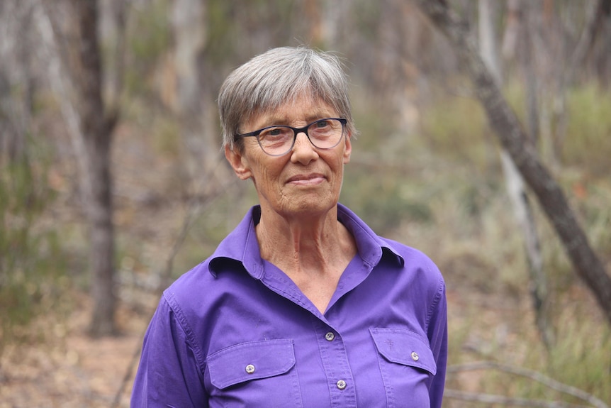 She has short grey hair and wears a bright purple shirt and stands among tall trees