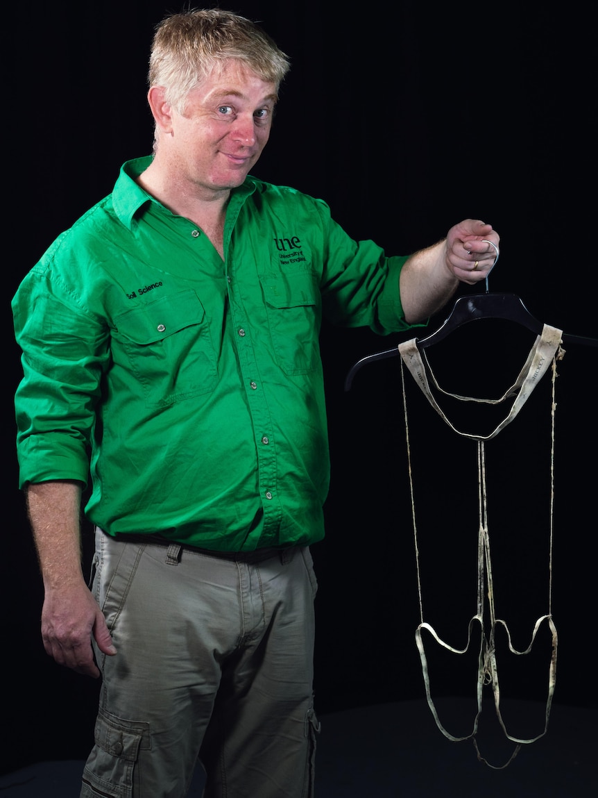 Young man with a pale complexion in a bright green embroidered work shirt holds a coat hanger with a pair of degraded long johns