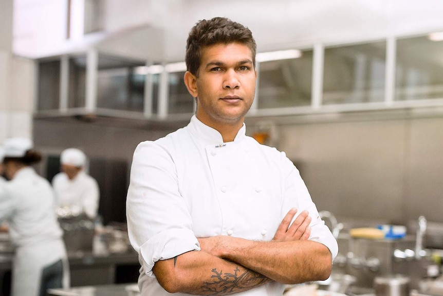 A young man wearing white chef jacket stands with arms crossed, behind him are two chefs working in stainless steel kitchen.