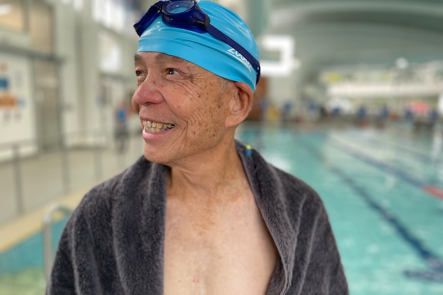A man in a swimming cap, goggles and a towel smiles from outside the pool 