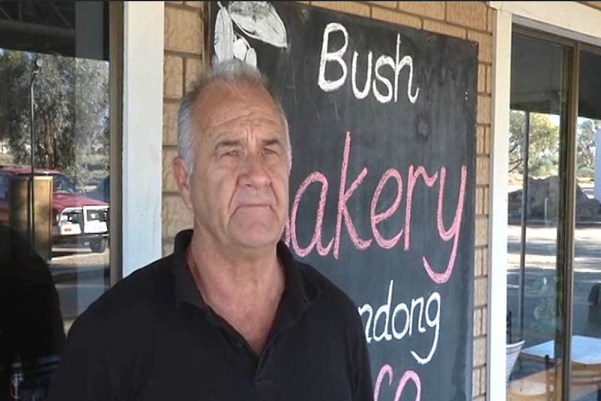 A man with greying hair stands out the front of a shop with a chalkboard in the background that reads "Bush Bakery"