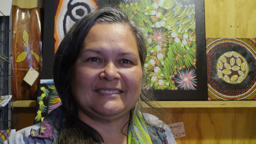 A woman stands in front of a shelf of paintings in Aboriginal style.