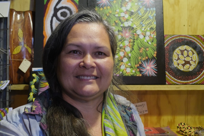 A woman stands in front of a shelf of paintings in Aboriginal style.