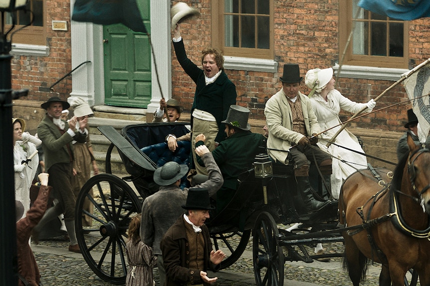 Colour still Rory Kinnear waving hat from horse-drawn carriage and addressing crowd in 2018 film Peterloo.