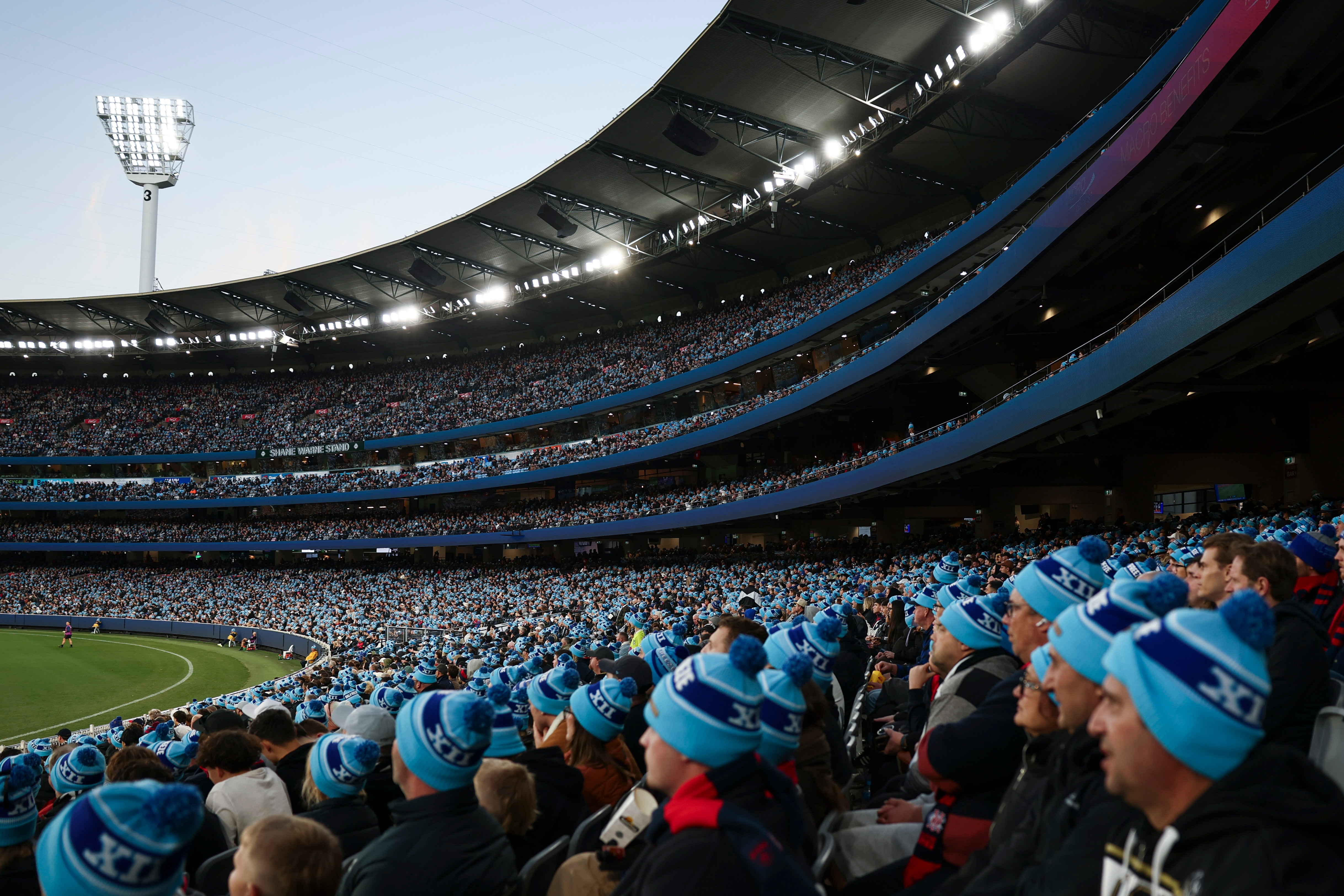 A large light pole stands over a packed sports stadium grandstand where nearly all the crowd is wearing a blue two-toned beanie.