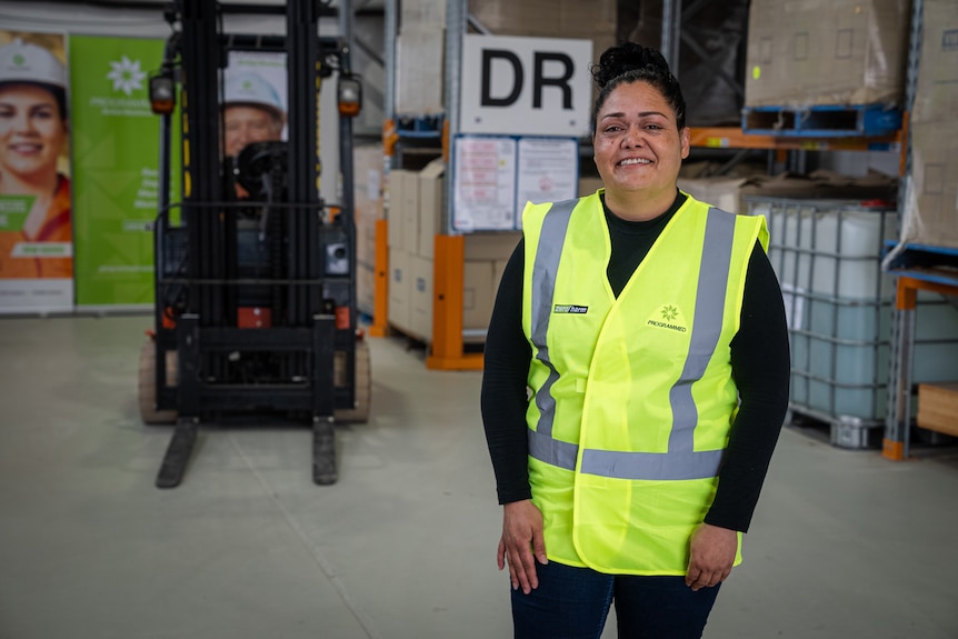 A women with her black hair tied up in a bun wears a bright yellow vest and stands in front of a forklift in a warehouse.