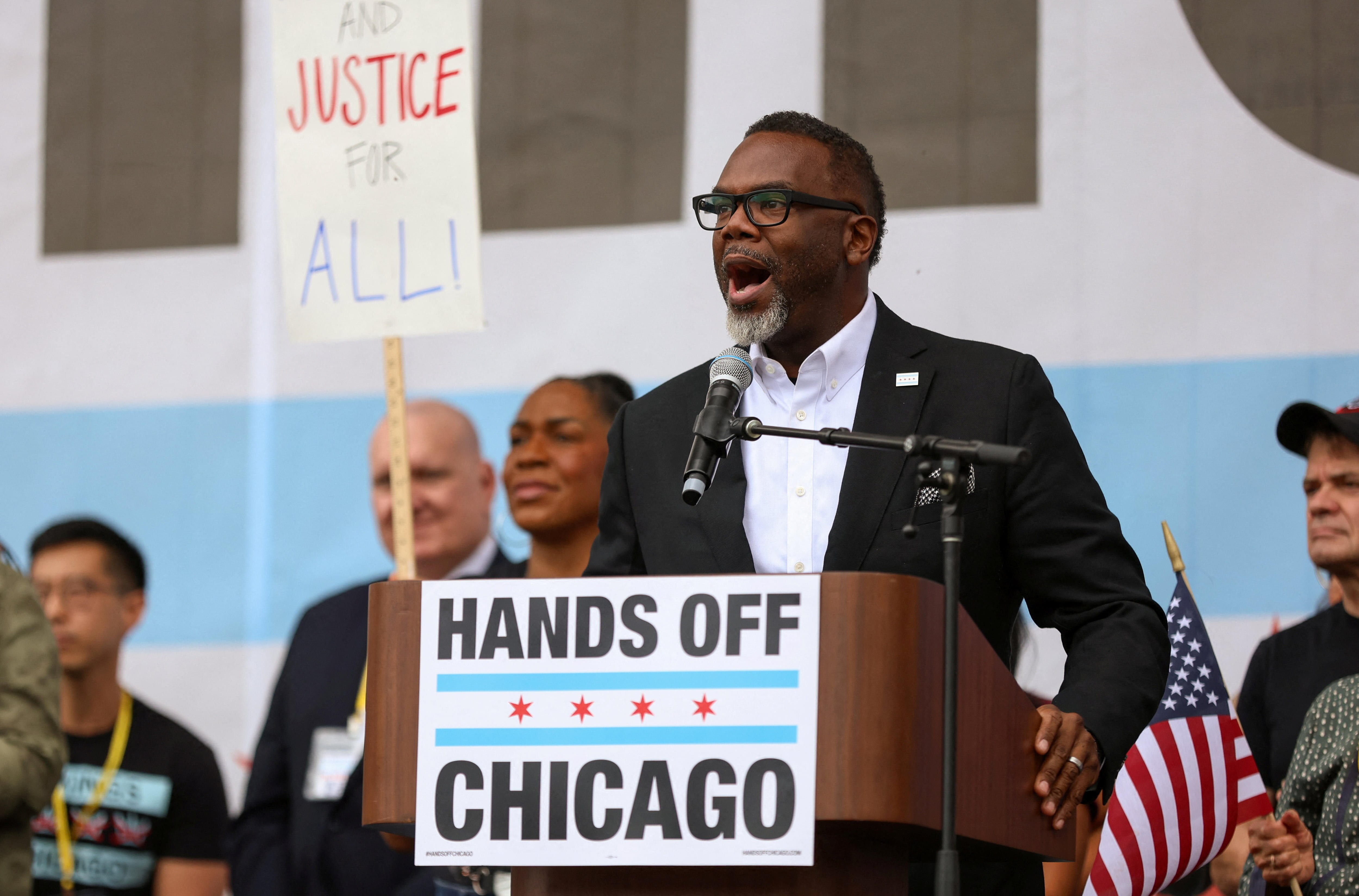 Brandon Johnson wearing a black suit and white shirt, speaking into a microphone at a lectern with a 'Hands Off Chicago' sign
