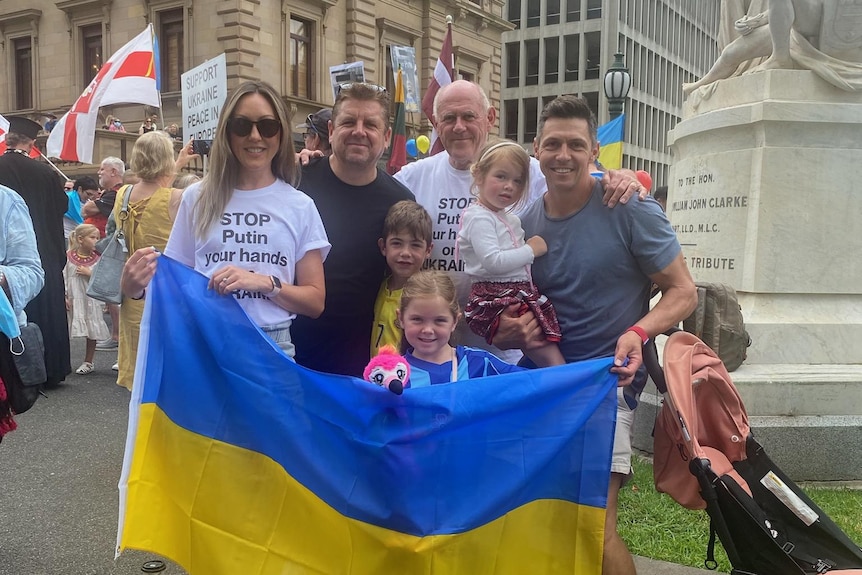 A family including to young children hold a Ukrainian flag in front of a monument at an outdoor rally.
