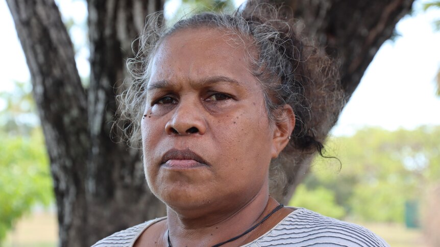 A woman looking directly at the camera looking sad and serious, standing outside with a tree in the background.
