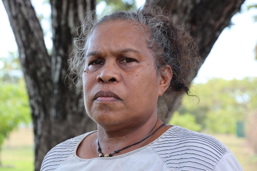 A woman looking directly at the camera looking sad and serious, standing outside with a tree in the background.
