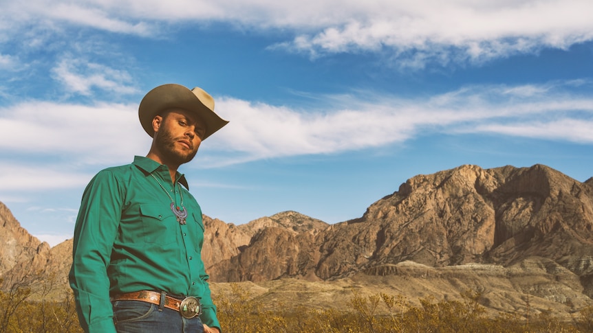 A man in a green shirt and 10 gallon hat looks down at the camera in front of picturesque mountains