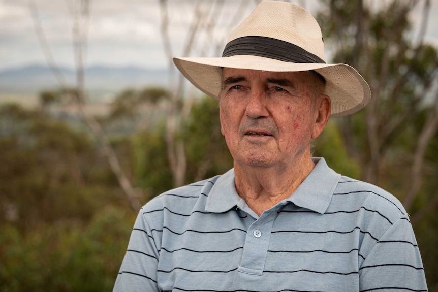 Rob McIlveen wears a blue t-shirt with stripes and a white hat, blurred out trees in the background.