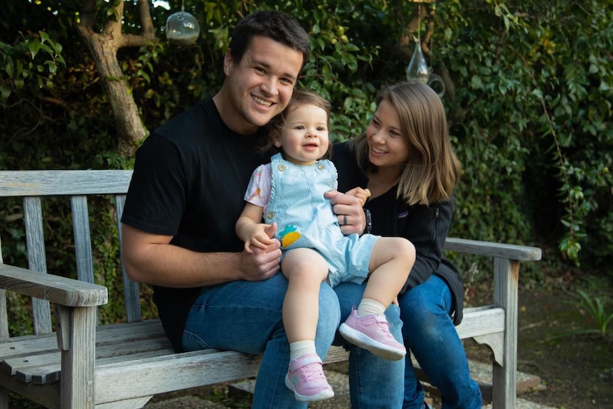 A smiling young family sitting on a park bench.
