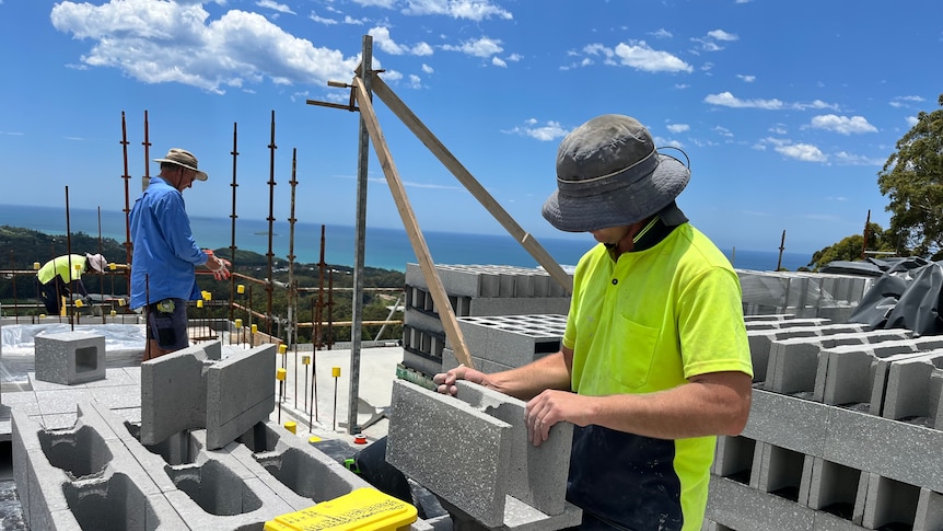 Lachie holds up a block on top of a stack of other blocks. Tony Crockett is seen working in the background.