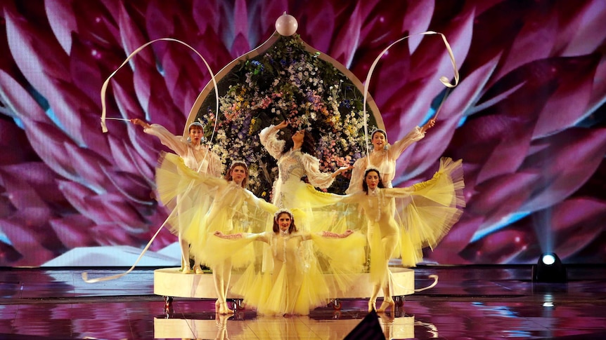 A Eurovision performer poses on a stage surrounded by backing singers and dancers.