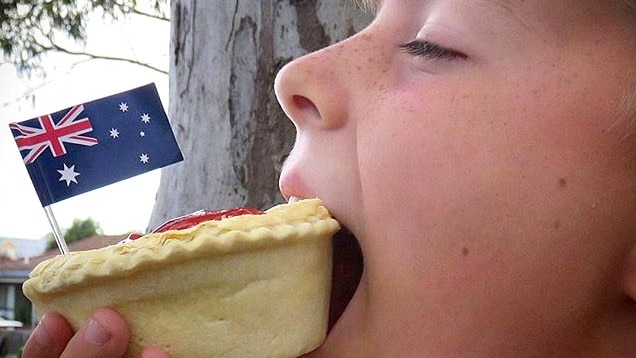 Close up, side on view of a boy about to bite into a meat pie, with a tiny Australian flag on it.