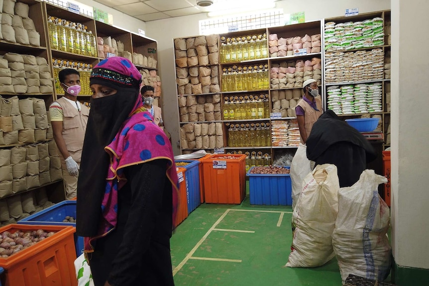 A Muslim woman with a pink headscarf and a cloth covering her face stands in front of shelves of cooking oil and bags of food.