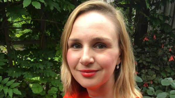 A woman with blonde hair and an orange top smiles at the camera in front of green leaves.