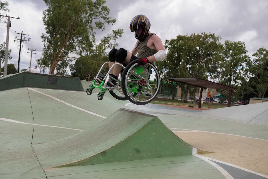 A man in a wheelchair gets air on a ramp. He is wearing a motorbike helmet and elbow guards.