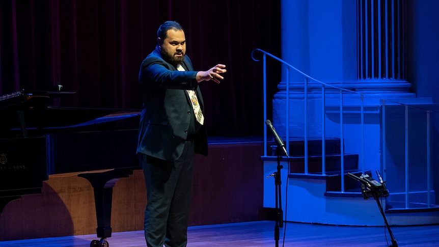 Tenor Manase Latu performs on stage. He has a serious expression and his arm is outstretched as if reaching for something.