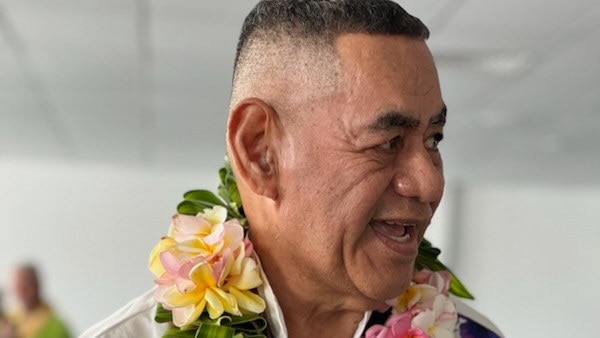Man wears white shirt with large brightly coloured flower lei around his neck. Looks to his left