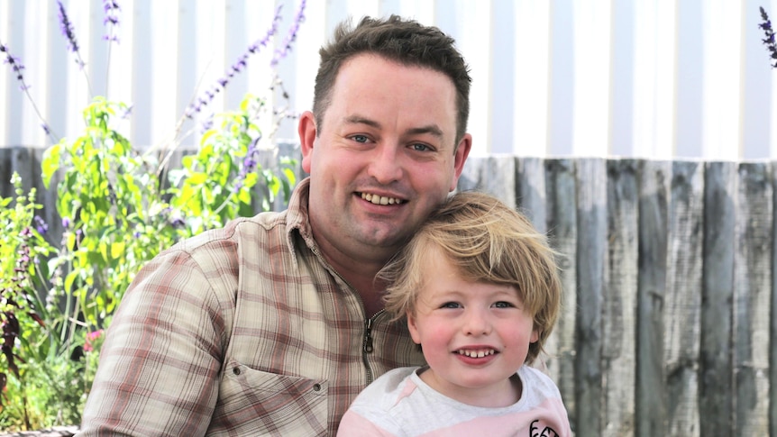 A man in a chequered shirt holds his son with shaggy blonde hair. Behind them is a fence and purple flowers.