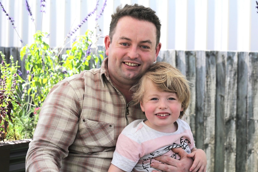 A man in a chequered shirt holds his son with shaggy blonde hair. Behind them is a fence and purple flowers.