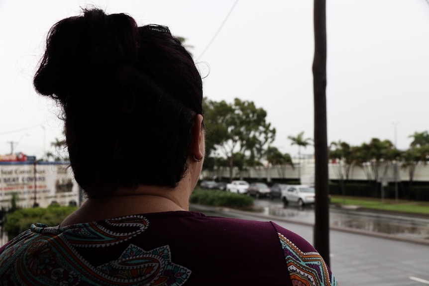 a woman stands with her back to the camera, as she watches a road near a mall