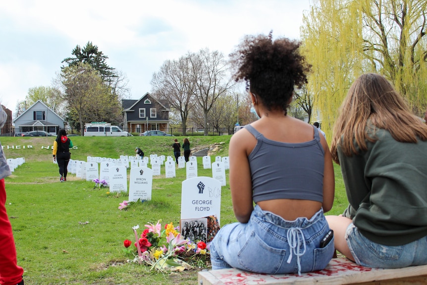 Two young women with their back to the camera look at rows of gravestones 
