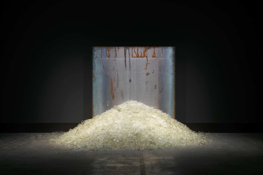 Dark room with spot-lit pile of broken glass, about two metres wide, in the middle, with a metal backdrop behind it.
