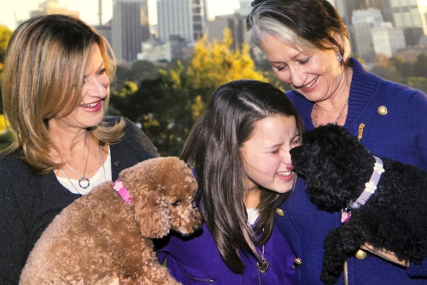 Two women smile as a black poodle licks a girl on the face