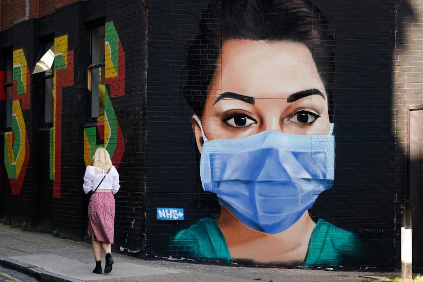 A woman walks past a mural on a building wall depicting a nurse in a mask