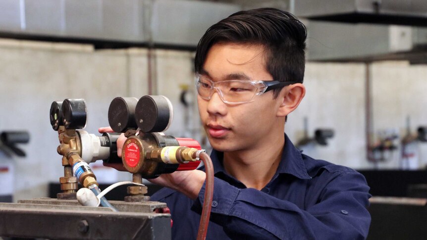 16-year-old Tim Lee wearing safety glasses and adjusting levels on equipment in a TAFE workshop.