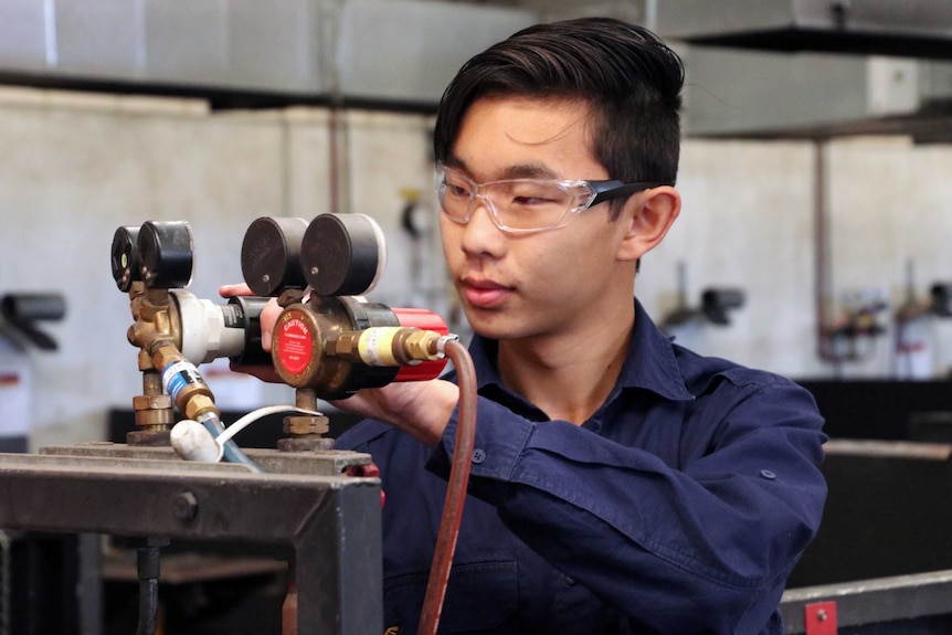 16-year-old Tim Lee wearing safety glasses and adjusting levels on equipment in a TAFE workshop.