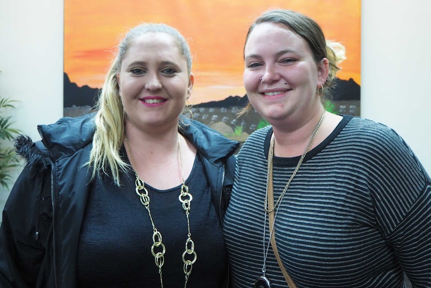 Two women stand together smiling in front of painting.