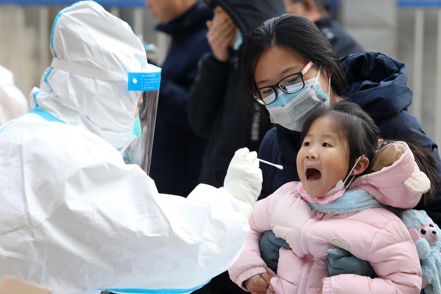 A young girl opens her mouth while sitting on a masked woman's lap, as a figure in PPE points a cotton swab at her.