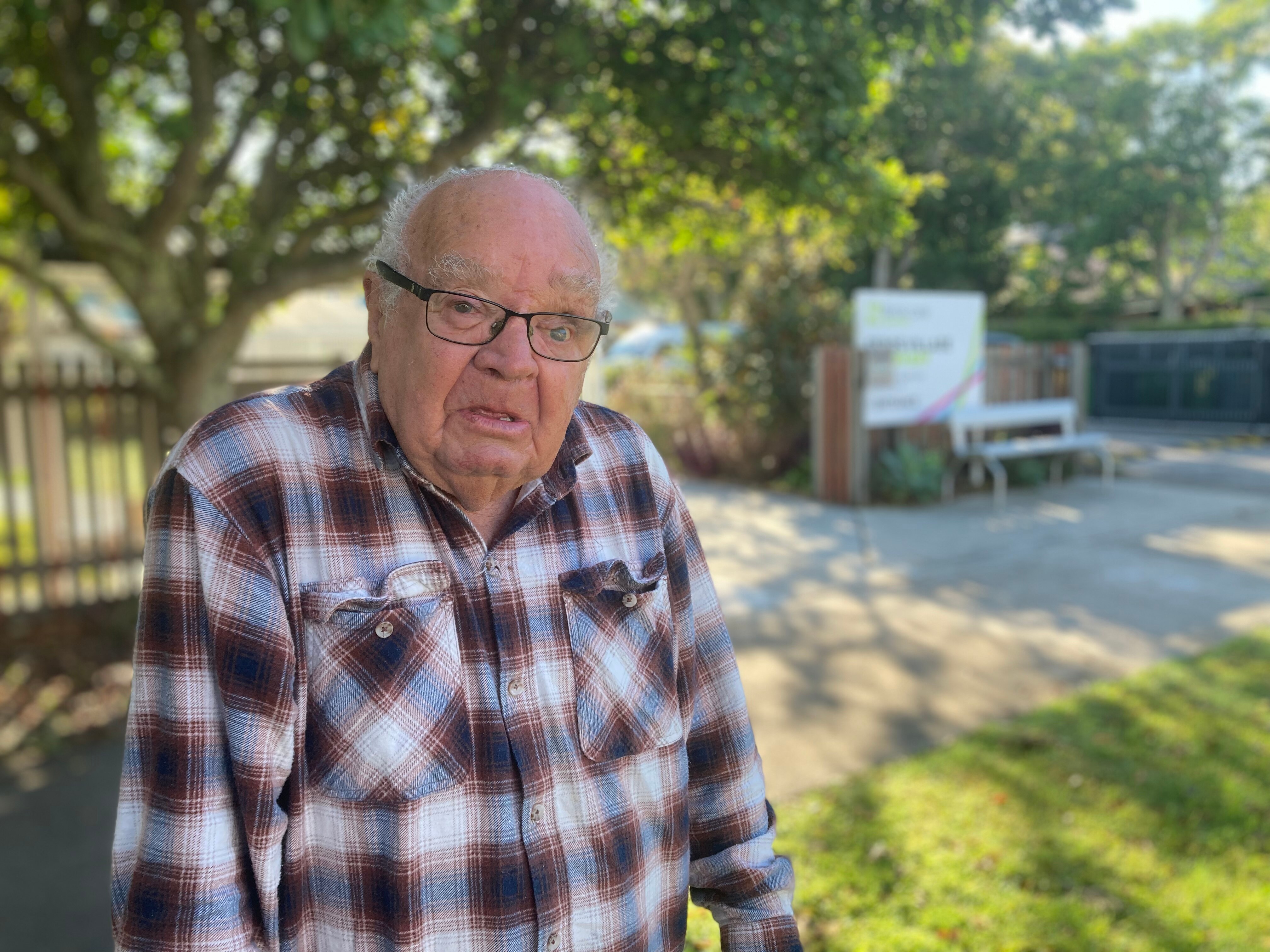 An elderly man in a checkered shirt and glasses stands outside a retirement home.