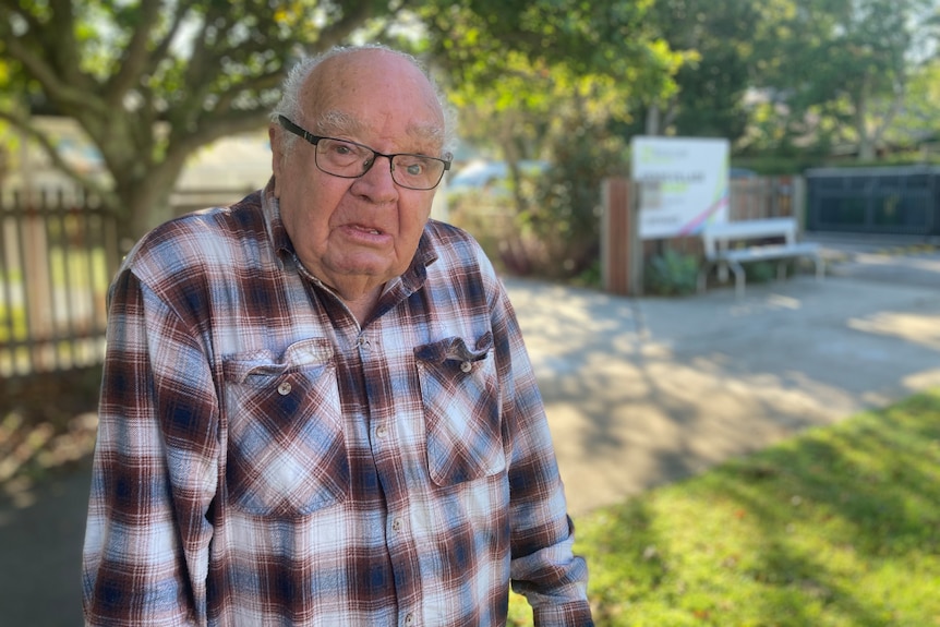 An elderly man in a checkered shirt and glasses stands outside a retirement home.