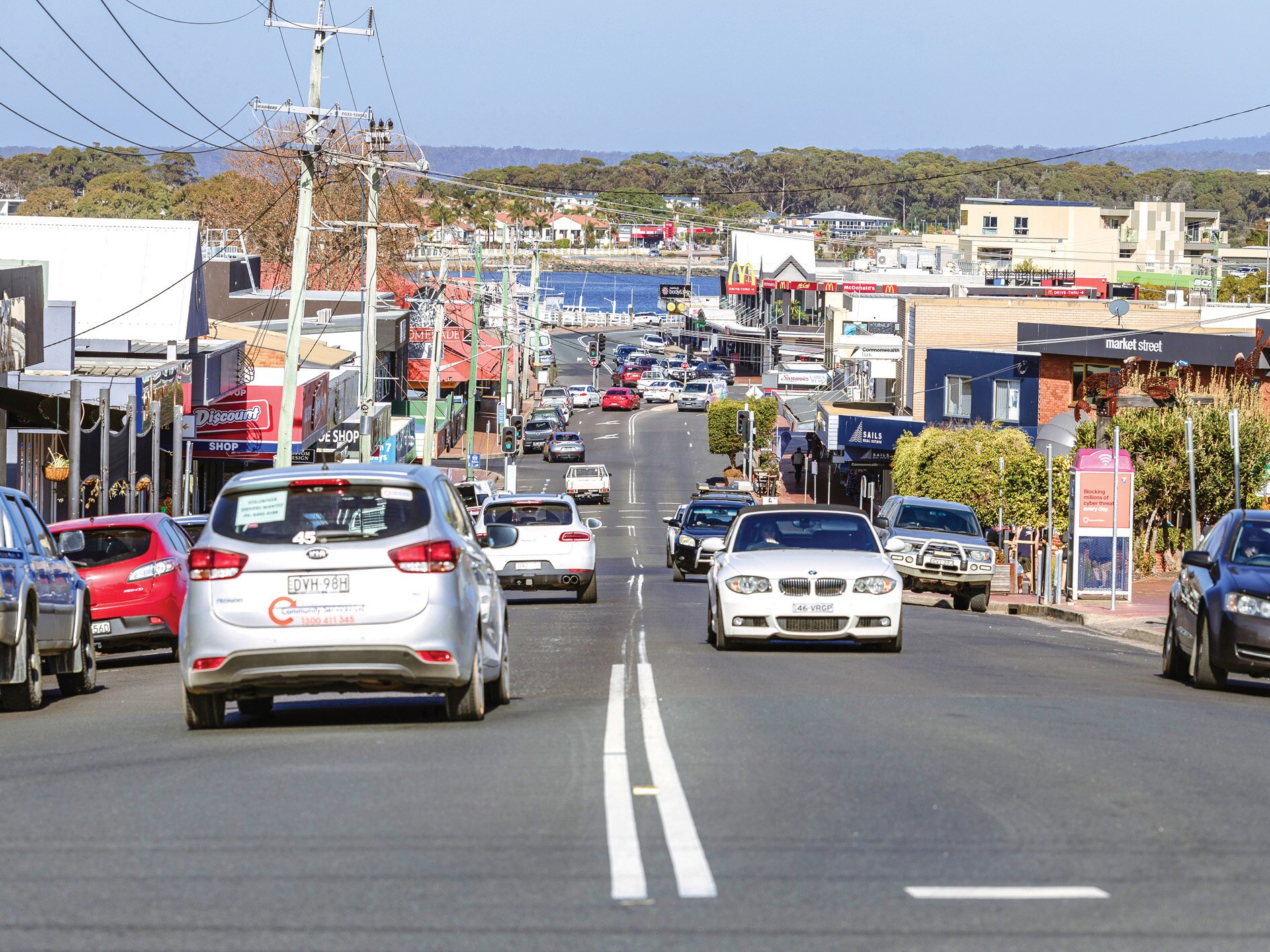 Main street of a country town with shops on either side and cars driving past, with water in the distance.