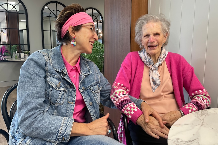 A woman with a pink headband and denim jacket sits next to and holds hands with an elderly woman in a pink cardigan. 