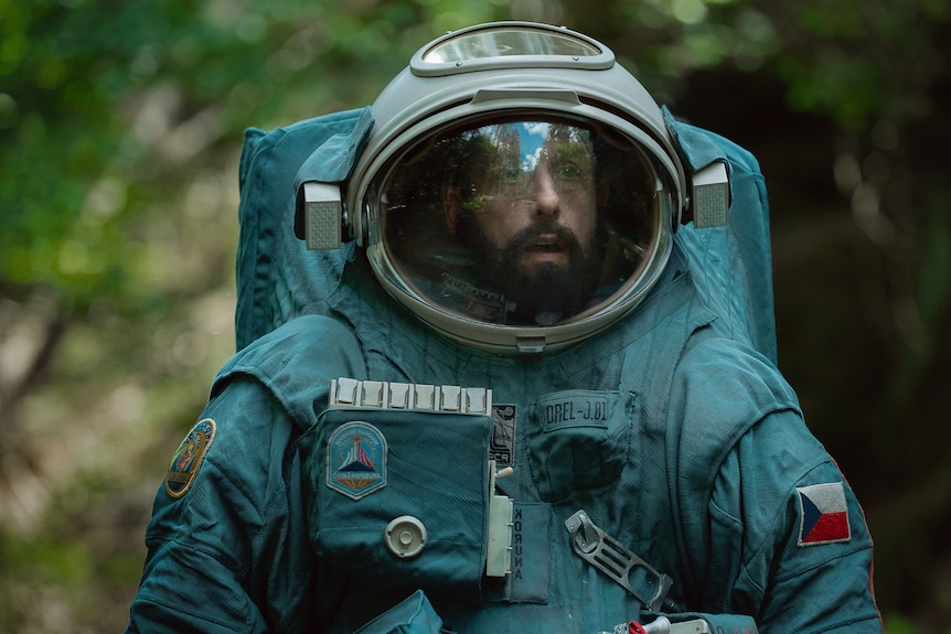 A film still showing a middle-aged man with a brown beard wearing an astronaut suit and helmet