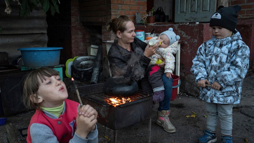 A child in the foreground blows on a burning stick while a woman feeds a baby next to a fire. Another child watches.