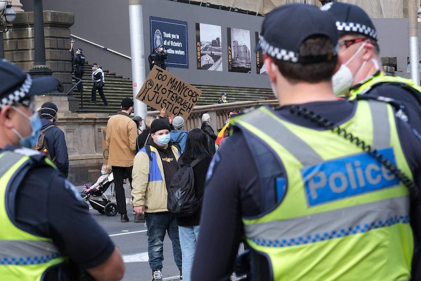 A protester holds a sign saying 'too many cops, not enough justice' in front of three police officers.