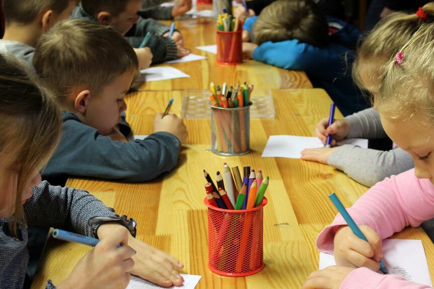 Children lined up along desks in kindergarten drawing on paper.