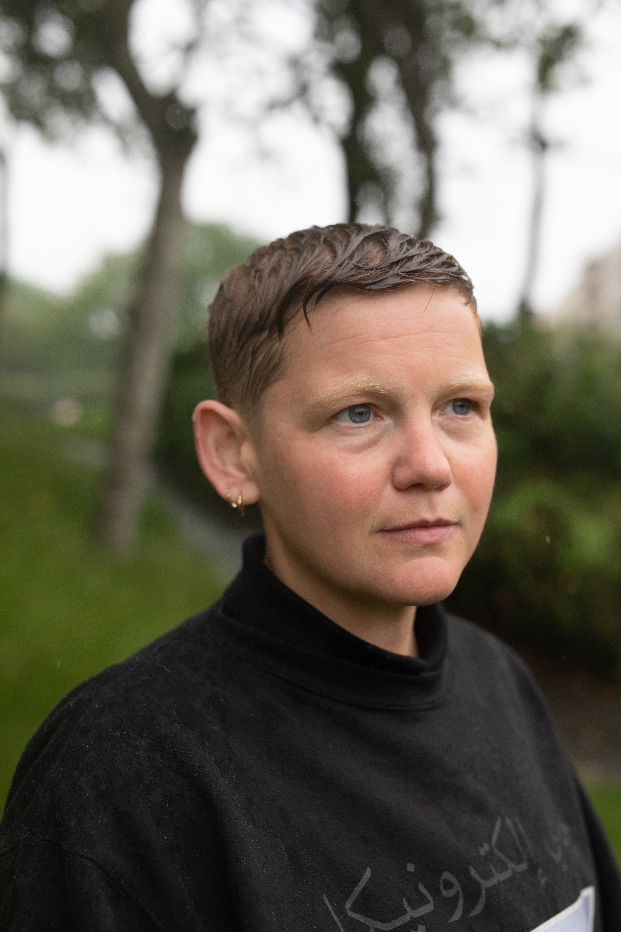 A white non-binary person with short sandy hair that is wet with rain stands in a park looking pensive.