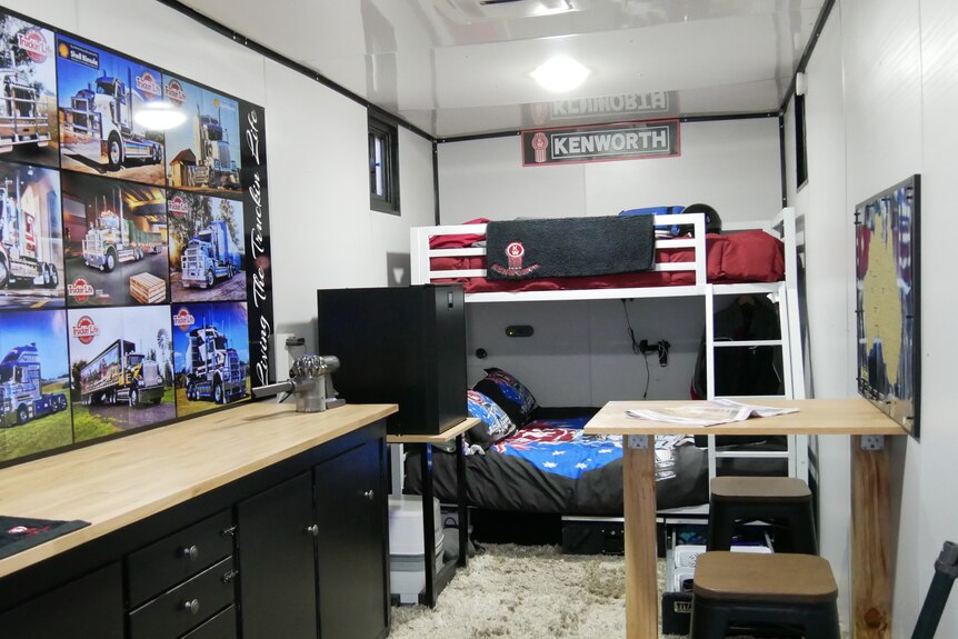 A room built inside a shipping container with a kitchenette, fridge, bunk beds, table and chairs and posters on the wall.